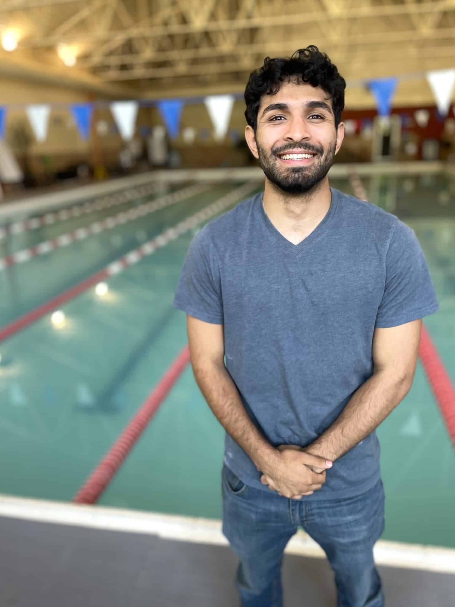 A man in a gray t-shirt and jeans stands smiling in front of an indoor swimming pool with lane dividers and flags visible in the background.