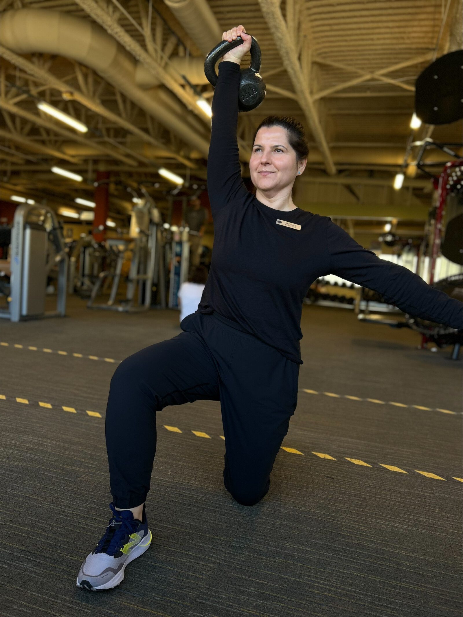 Certified trainer kneeling on one knee in black athletic wear, holding a kettlebell overhead in a gym setting. West Seattle Personal Trainers trust the expertise of professionals certified by the National Academy of Sports Medicine.