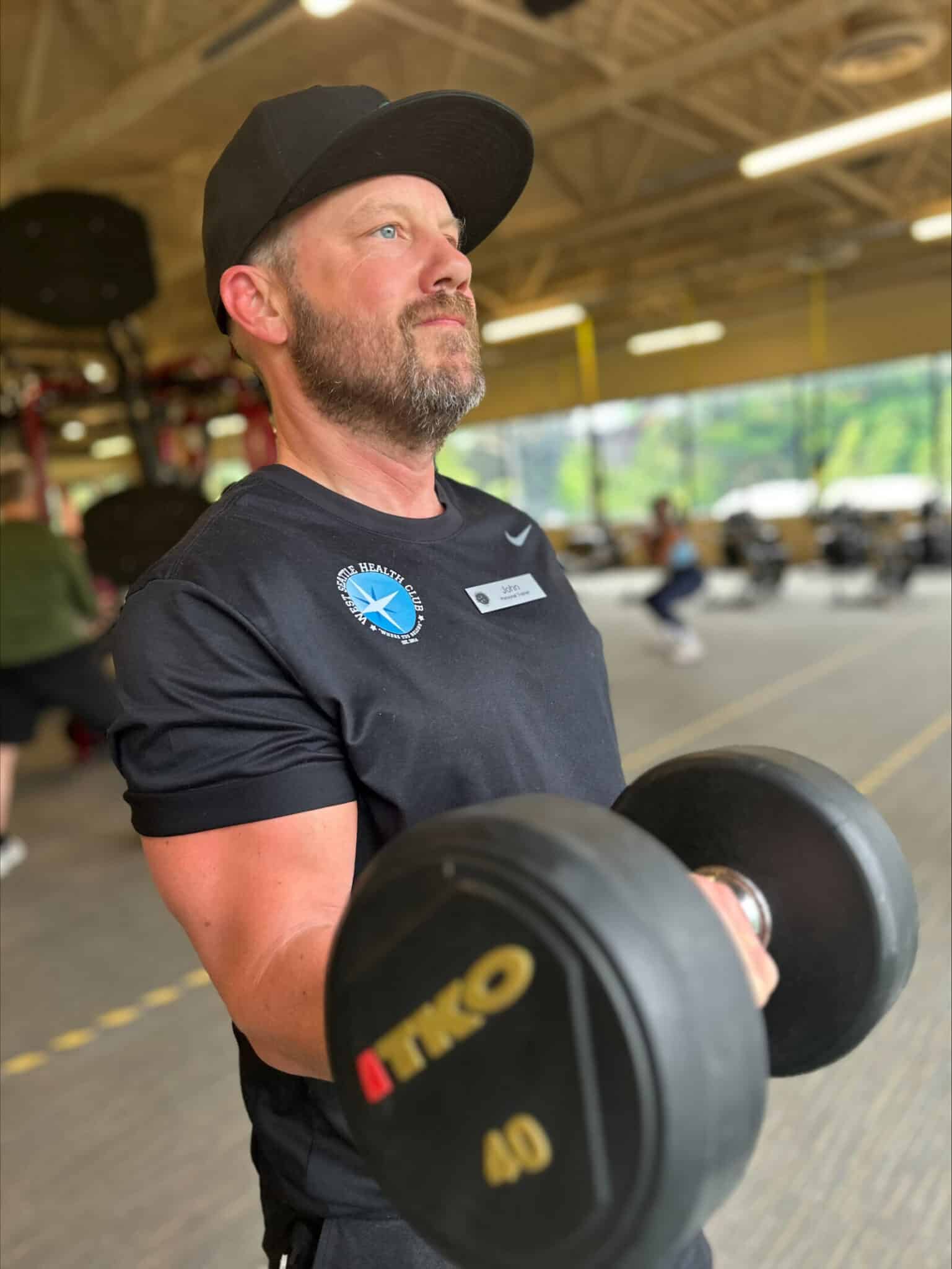 Man wearing a black shirt and cap lifts a 40-pound dumbbell in a gym, showcasing his strength under the guidance of a certified trainer from West Seattle Personal Trainers, with equipment and others visible in the background.
