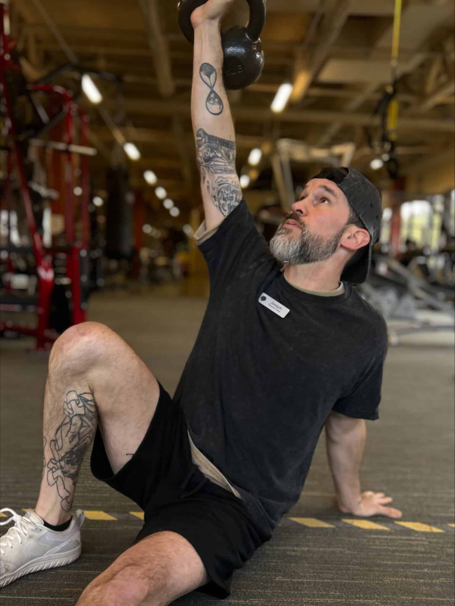 A man wearing a black t-shirt and shorts does a Turkish get-up with a kettlebell in a gym, guided by a certified trainer from the National Academy of Sports Medicine, looking upward with one arm extended.
