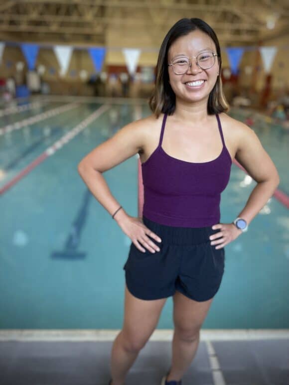 A woman in athletic wear stands and smiles beside an indoor swimming pool, with swim lanes and banners visible in the background—a proud member of the West Seattle Swim Coaches team.