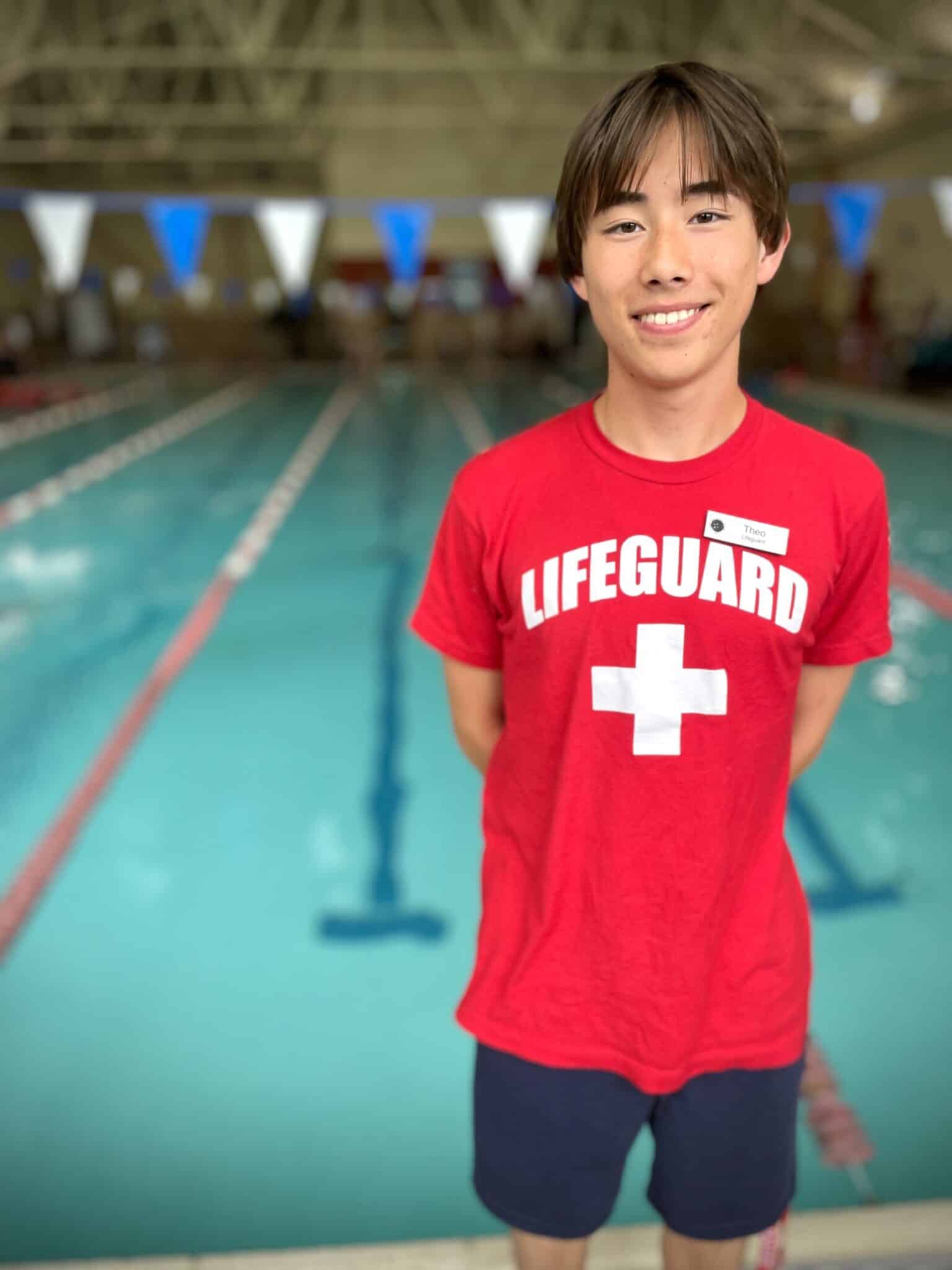 A teenage lifeguard in a red shirt stands at the edge of an indoor swimming pool, smiling at the camera—ready to assist swimmers and West Seattle Swim Coaches.