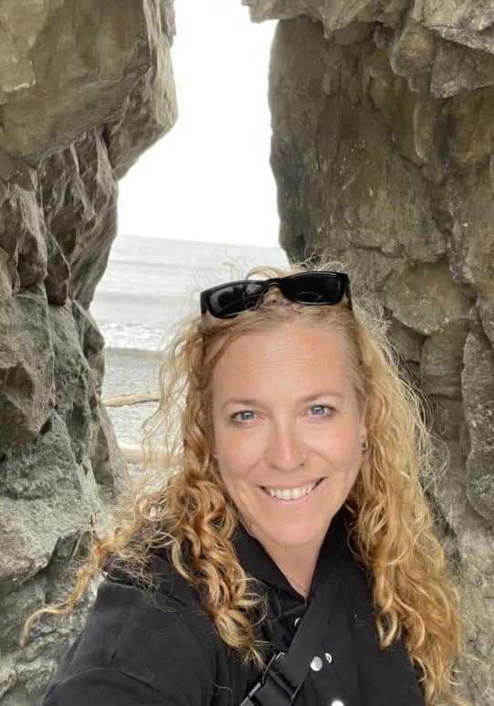 A woman with curly blonde hair and sunglasses on her head stands between large rocks, smiling at the camera with the sea visible in the background.