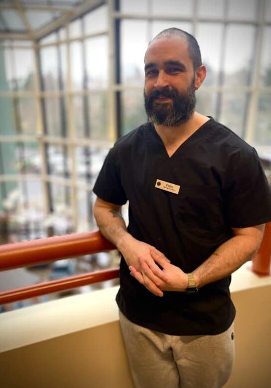 A man with a beard wearing black scrubs and a name tag stands indoors by a red railing, with large windows and an outdoor view in the background.