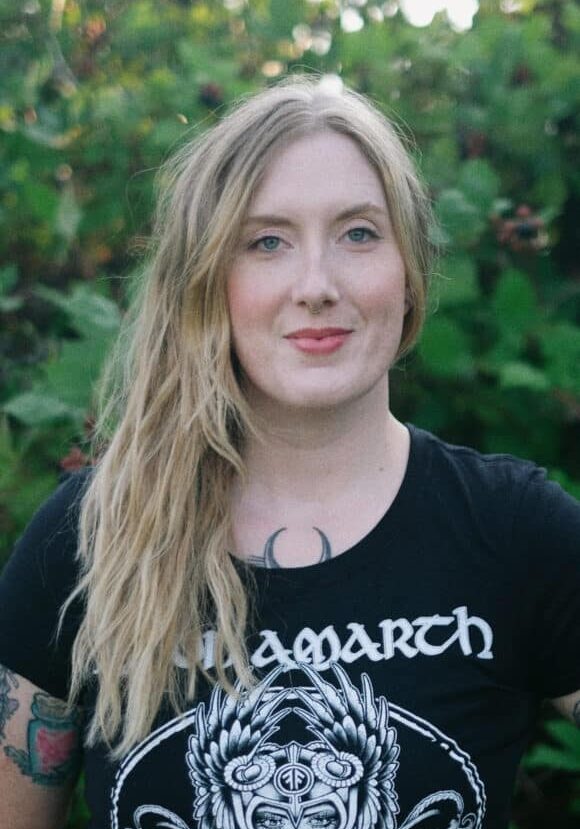 A woman with long blonde hair stands outdoors in front of green foliage, wearing a black graphic T-shirt and showing a crescent moon tattoo on her chest.