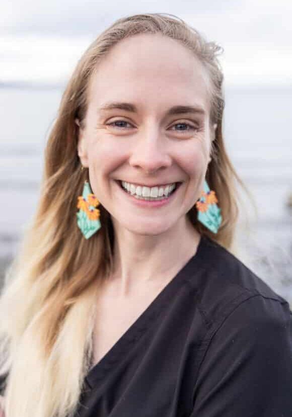 A woman with long blonde hair smiles at the camera, wearing colorful flower earrings and a black top, with a blurred outdoor background.