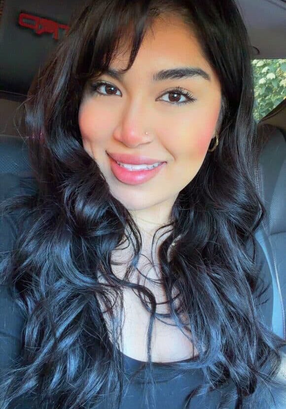 A woman with long wavy dark hair and a nose piercing smiles while sitting in a car, wearing a black top.