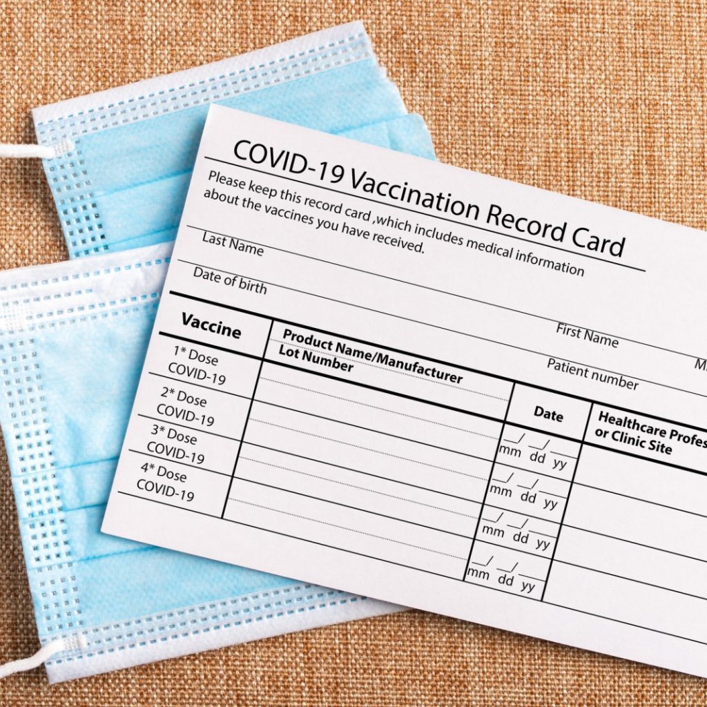 A COVID-19 vaccination record card rests atop two blue disposable face masks on a burlap surface, echoing the recent Covid updates at West Seattle Health Club.