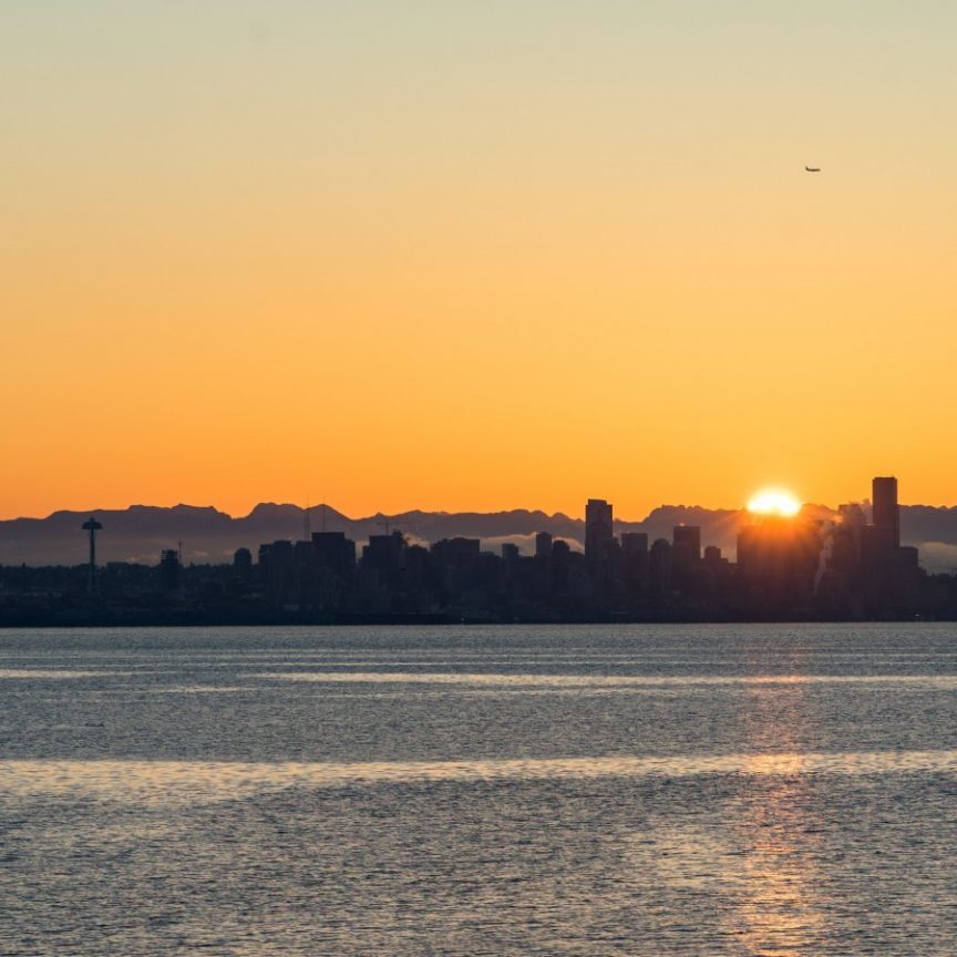 Silhouette of a city skyline at sunset with an orange sky, mountains in the background, and a body of water in the foreground; proof of vaccination no longer required for entry into this serene oasis.